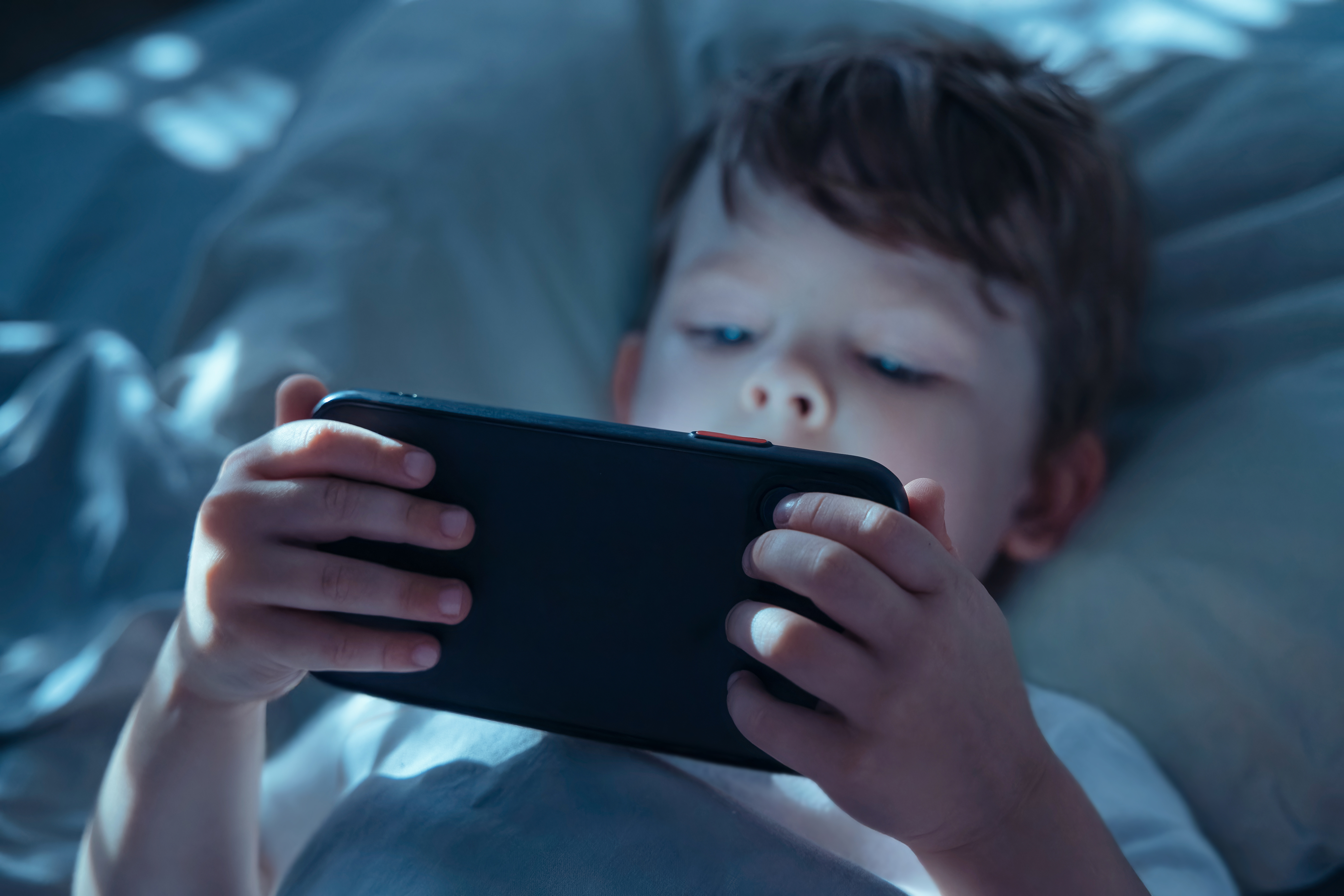 Little child lying in bed in dark room, holding smartphone close and watching intently, illuminated by blue screen light. Gadget addiction and screen dependency. Selective focus on smartphone. By Arsenii