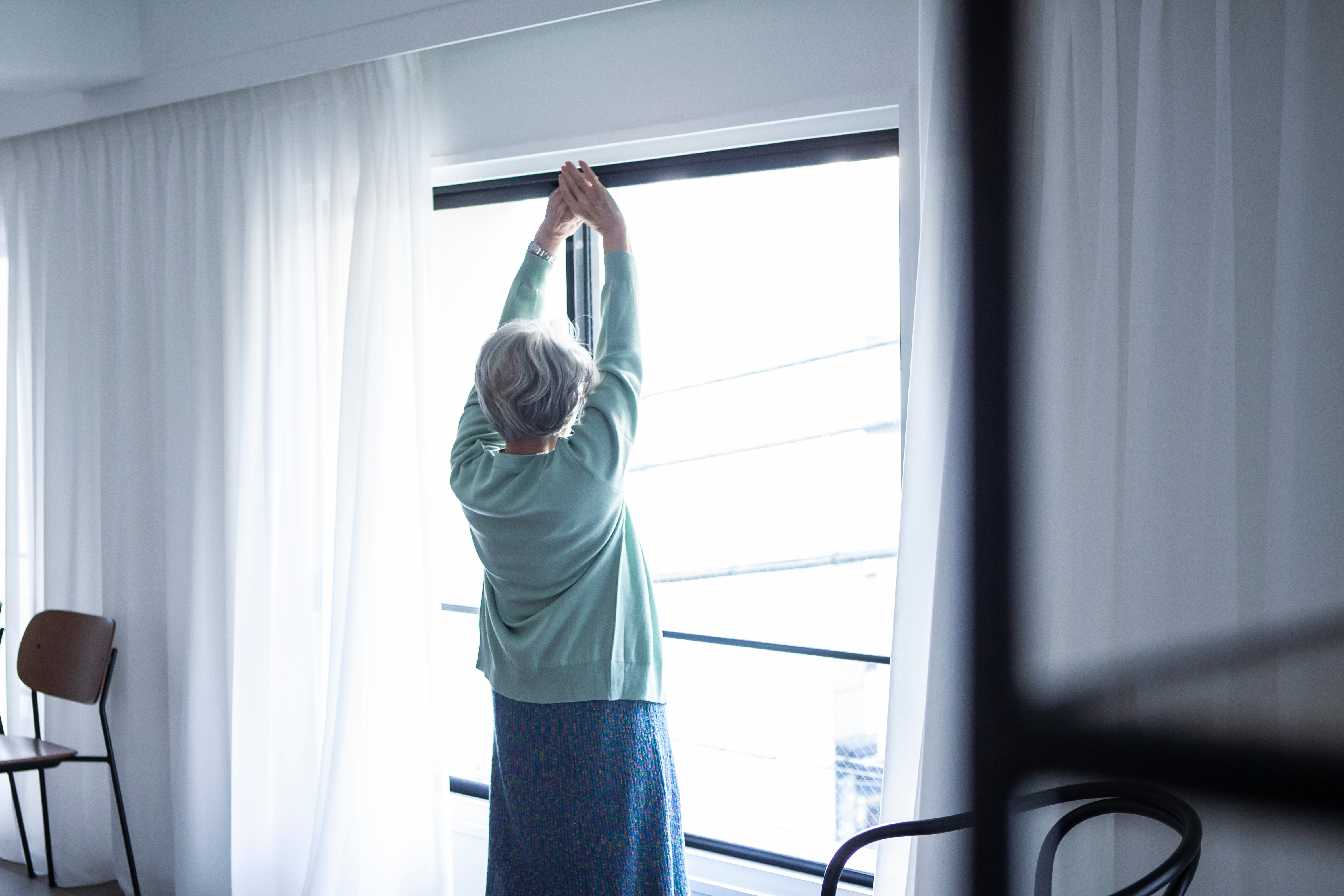 窓の外から朝日を浴びてストレッチするシニア女性 By Monet