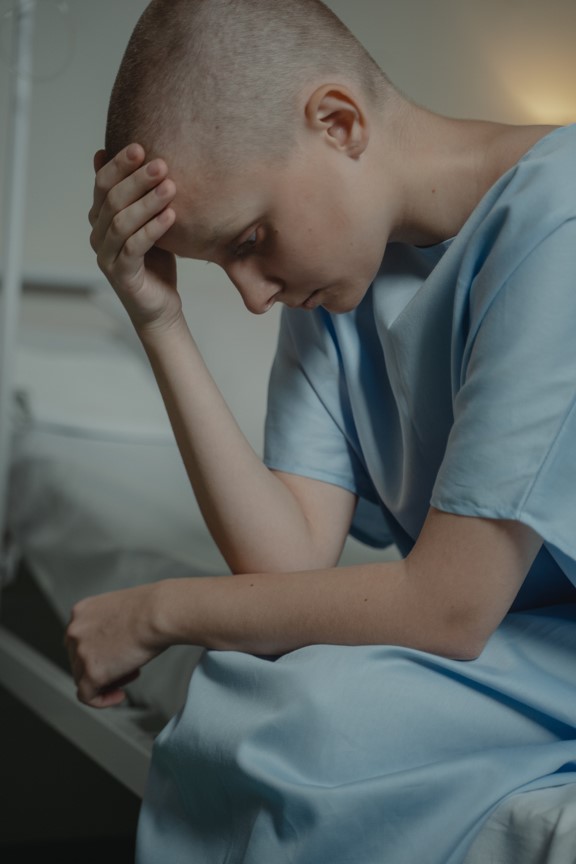 Bald woman in a hospital gown, sitting and looking down, symbolizes illness and contemplation. Copyright: Tima Miroshnichenko, Pexels.