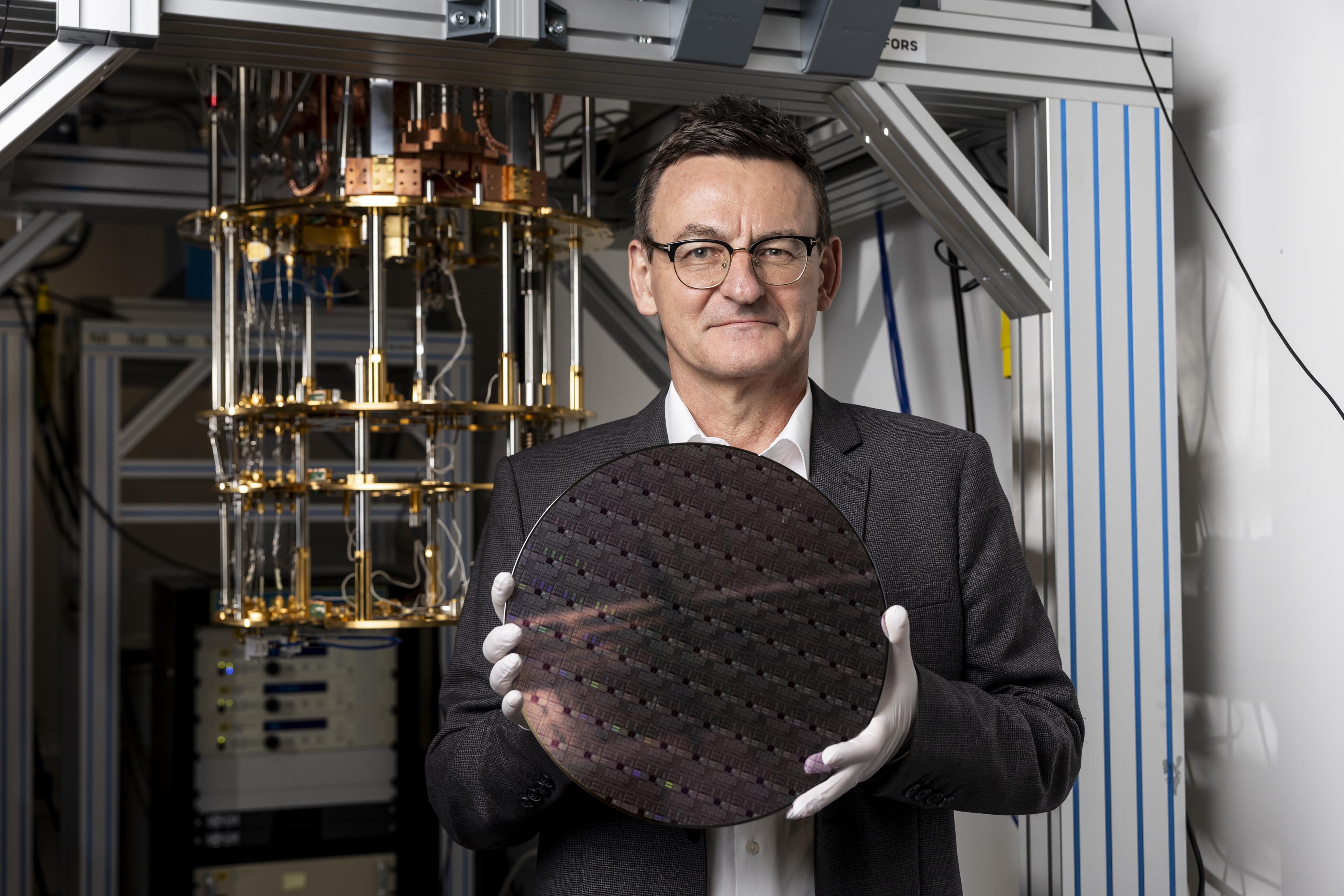 Diraq CEO and Founder, Andrew Dzurak, holding a 300 mm silicon wafer containing Diraq’s quantum computer chips, which were manufactured at Interuniversity Microelectronics Centre (imec) using standard semiconductor processes. The dilution refrigerator in the background was used to show that these chips achieve more than 99% fidelity in all quantum operations, surpassing the threshold required for quantum error correction. Credit: Diraq 